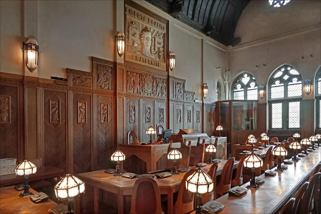 La salle du conseil municipal d'un hôtel de ville, vide : bancs de bois, lampes allumées, Marianne au mur.